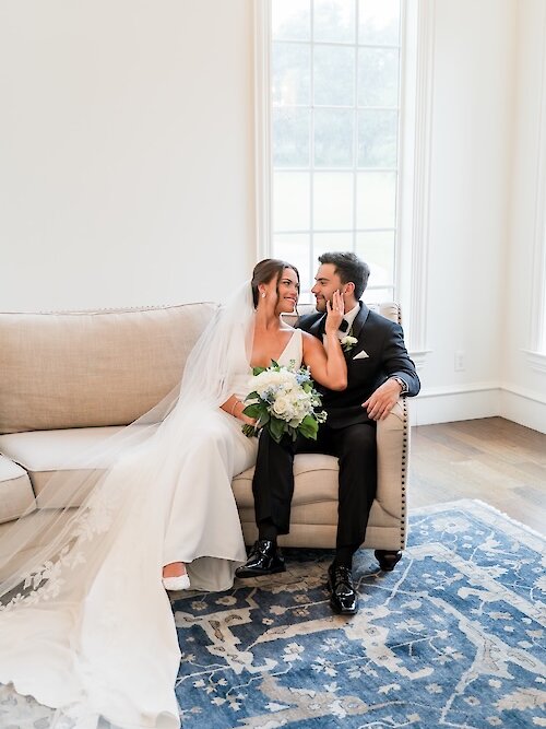 Bride and groom sitting together on a sofa, smiling and touching each other’s faces during an intimate indoor wedding portrait.