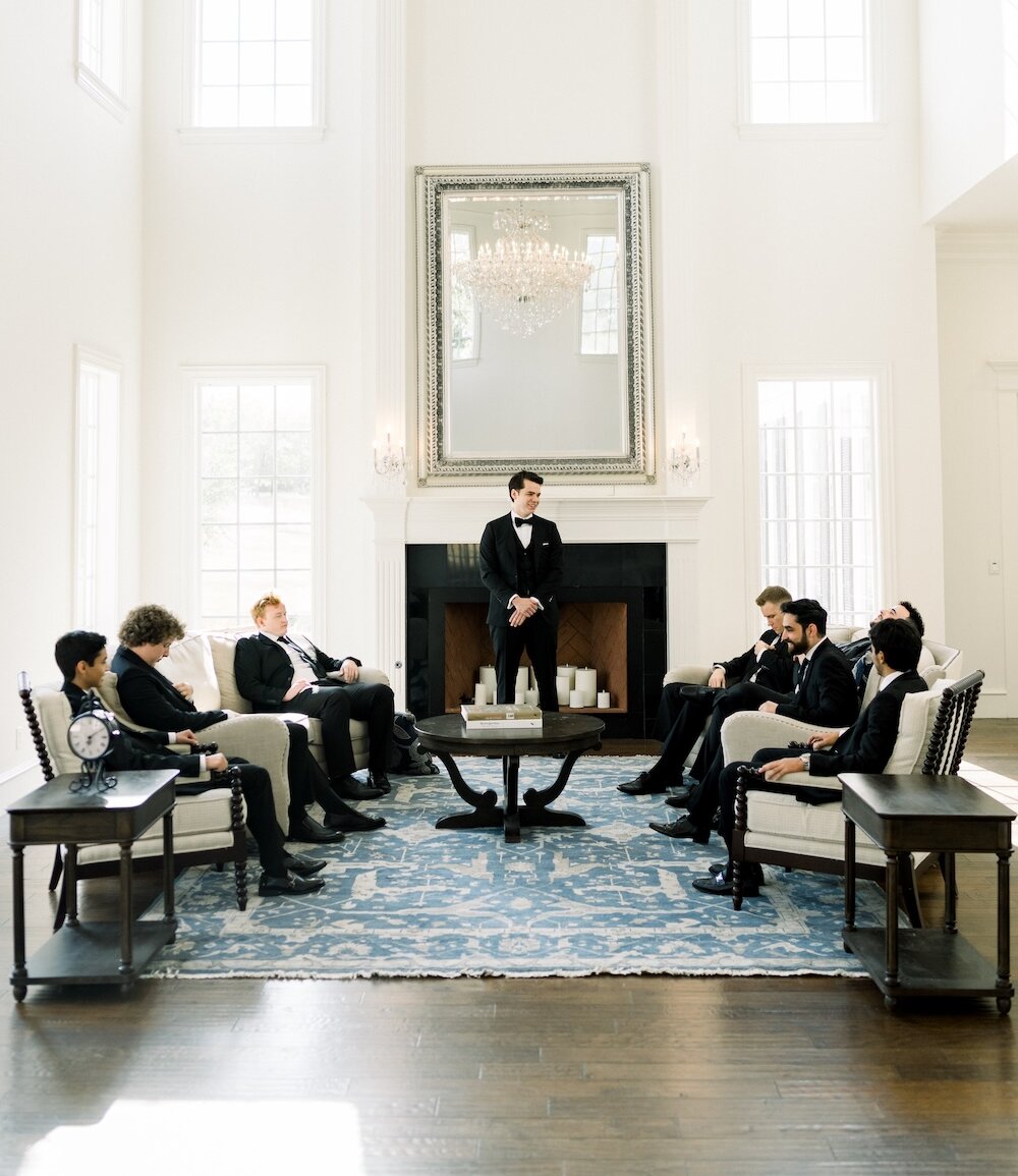 Groom standing in an elegant, bright sitting room with his groomsmen seated around him before the wedding ceremony.