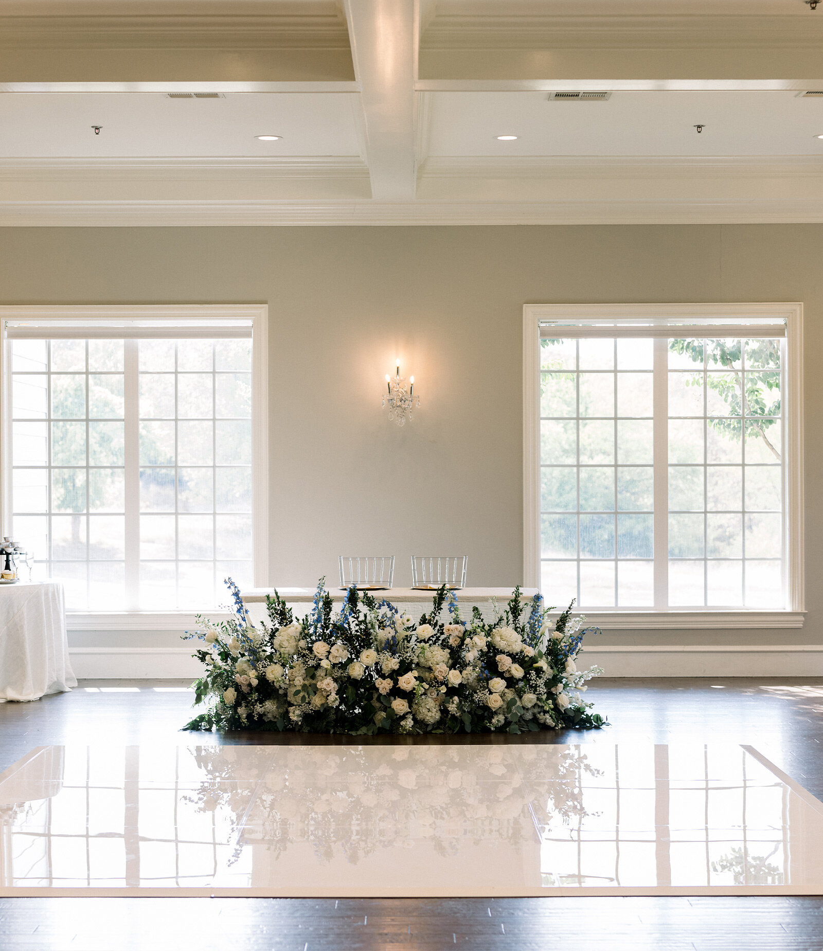 Elegant wedding reception setup with crystal chandeliers, clear chairs, white linens, and floral centerpieces in a bright ballroom.