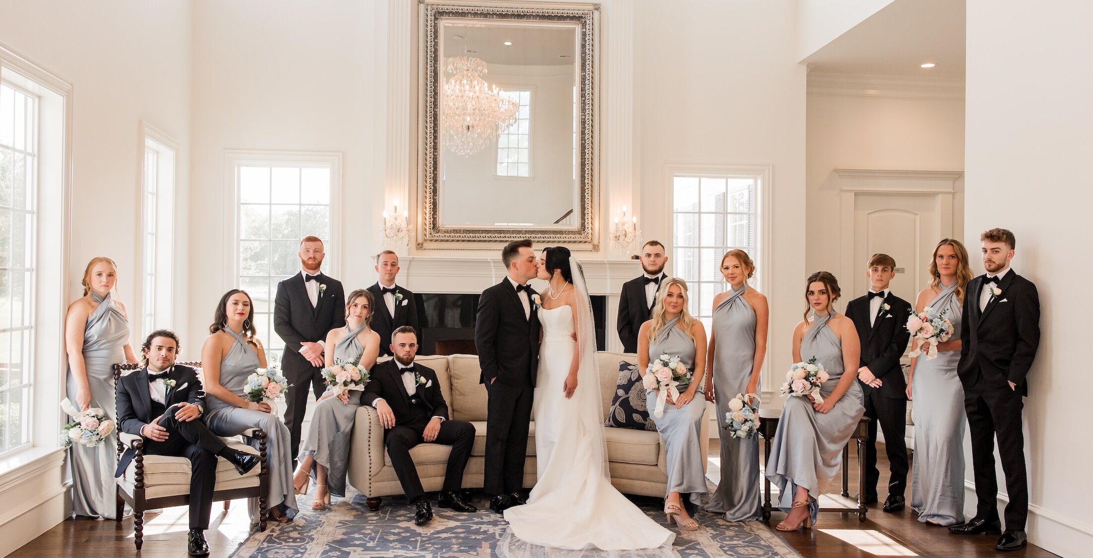 Bride and groom kissing in the center of their wedding party inside a bright, elegant room with tall windows and a crystal chandelier.