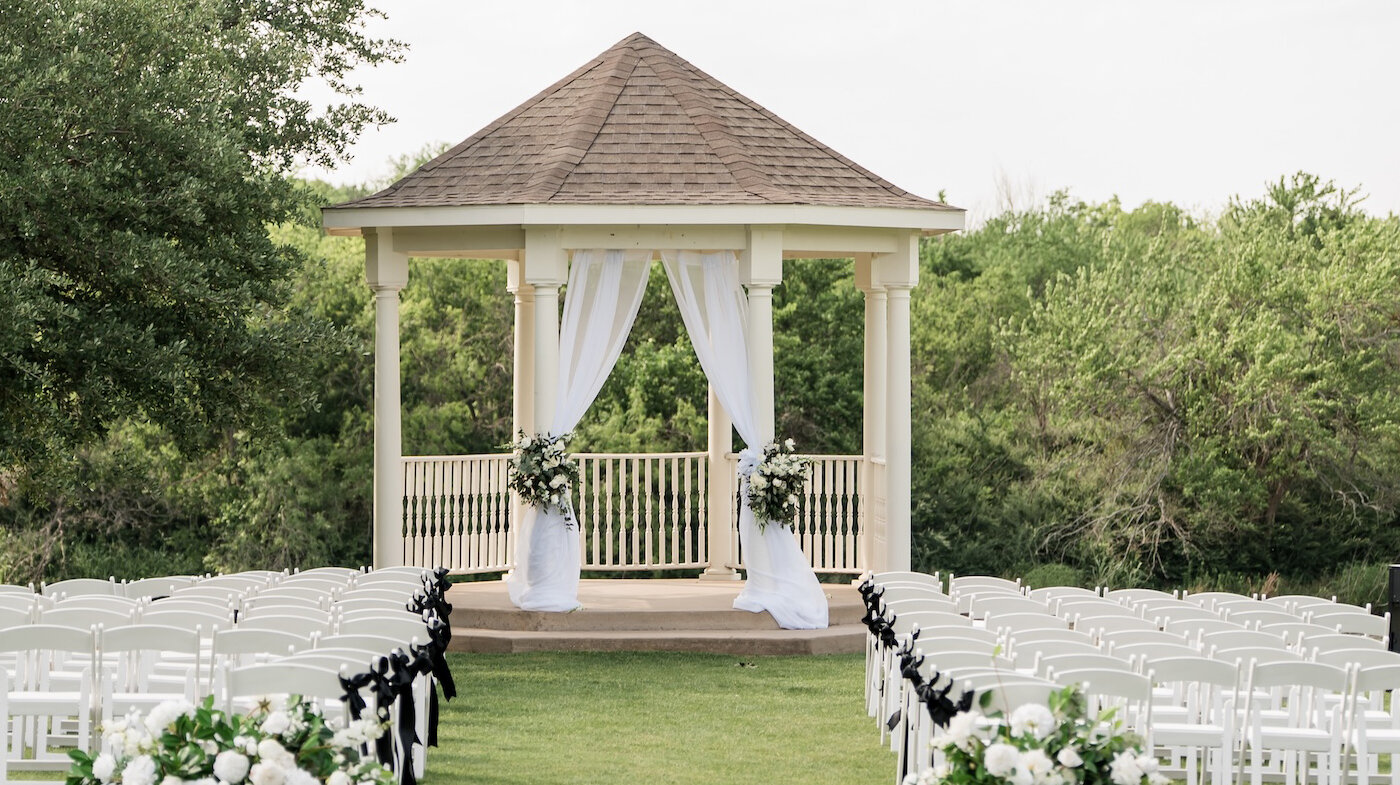 Outdoor wedding ceremony setup with white chairs, black ribbon accents, lush white floral arrangements, and a gazebo draped with white fabric surrounded by greenery.