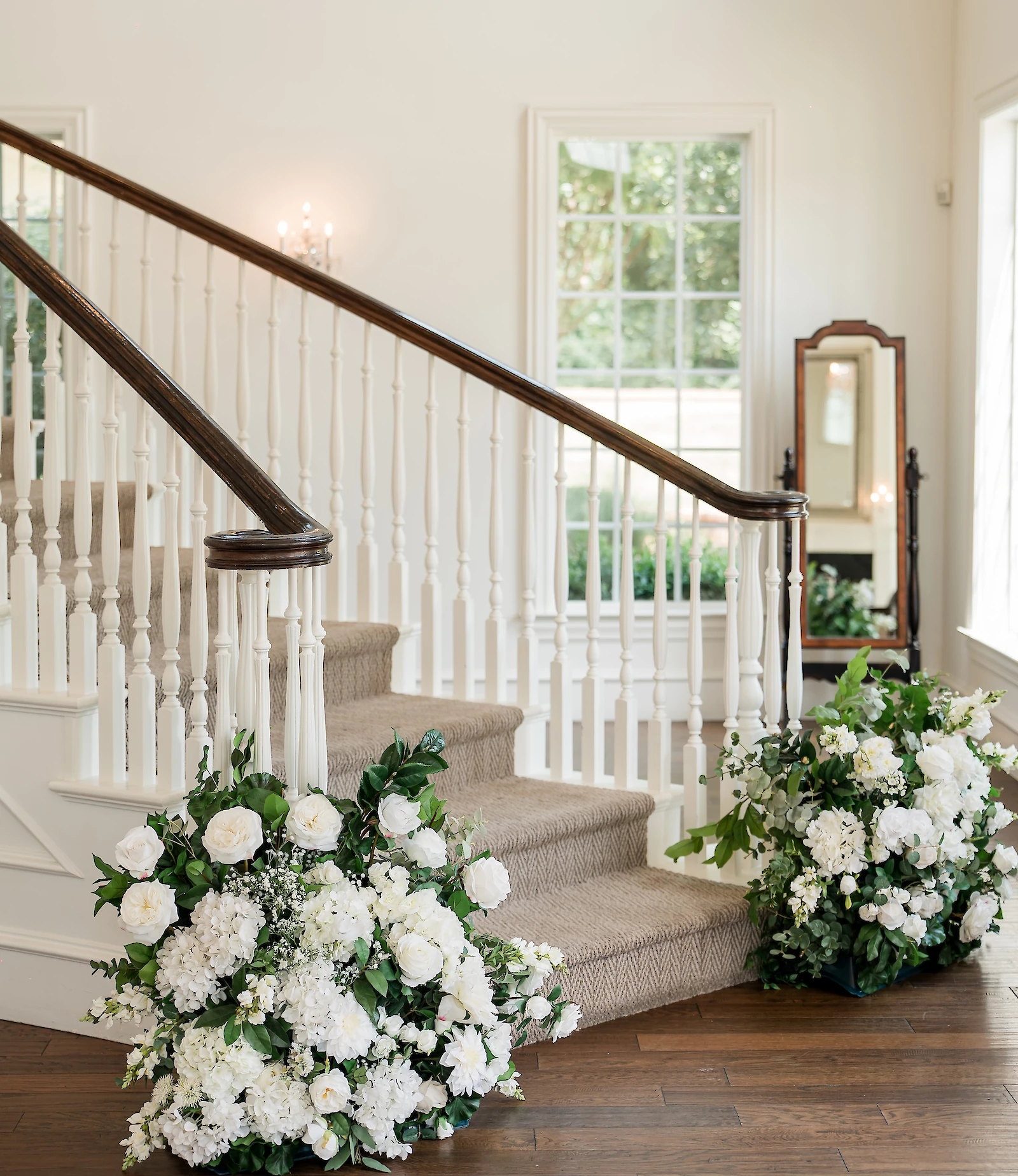 Staircase decorated with lush white floral arrangements inside a bright wedding venue with large windows and a full-length mirror.