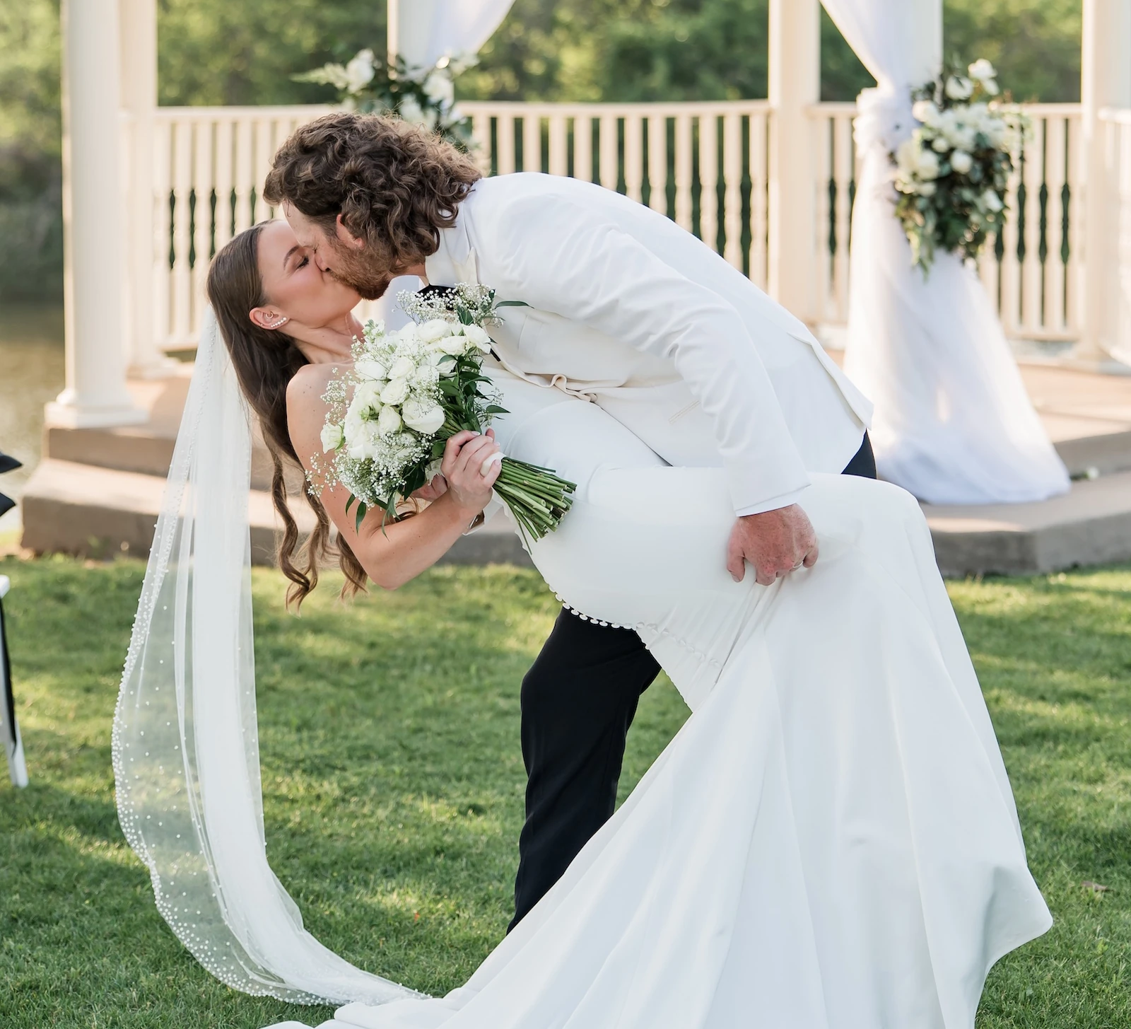 Groom dipping and kissing the bride in front of a white gazebo draped with fabric during an outdoor lakeside wedding ceremony.