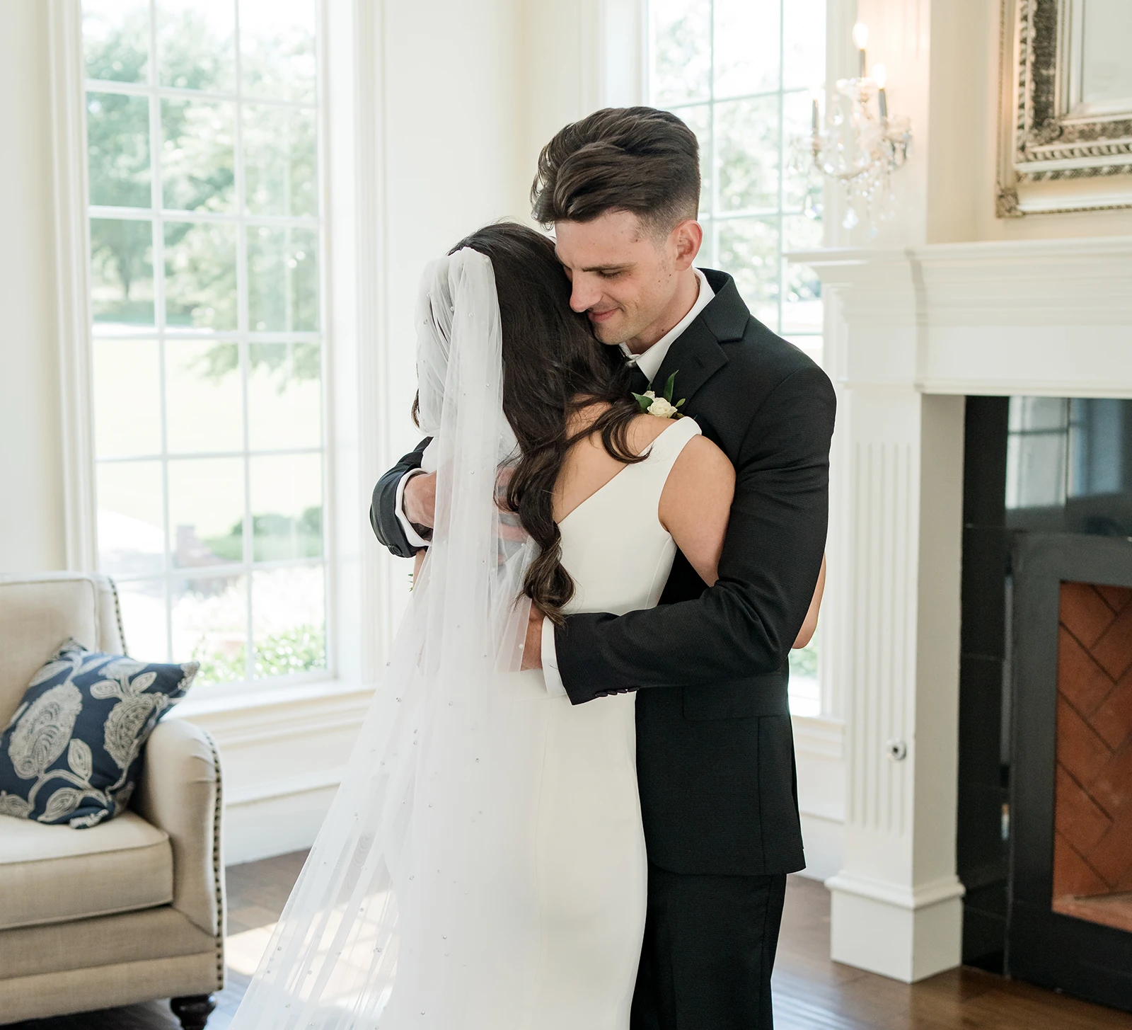 Groom and bride share an emotional first-look hug inside a bright room with large windows and elegant white decor