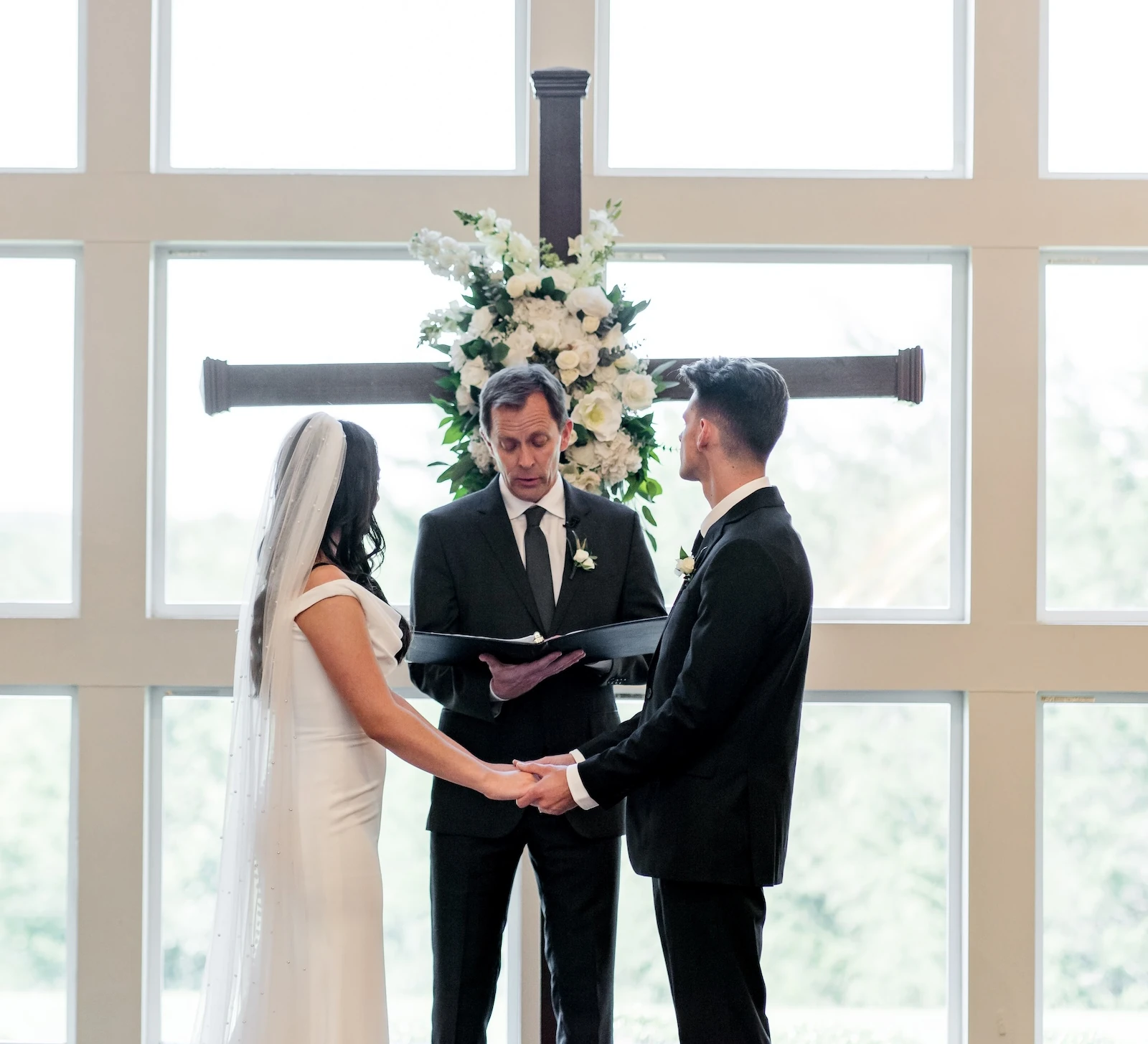 Bride and groom holding hands during their indoor wedding ceremony in front of a large cross and bright grid-pane windows.