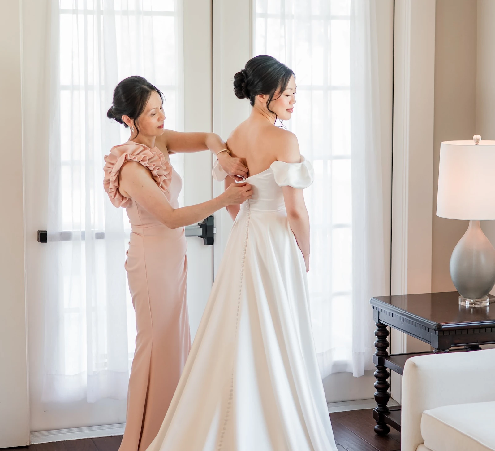 Mother helping the bride button her off-the-shoulder wedding gown in a softly lit room with French doors.
