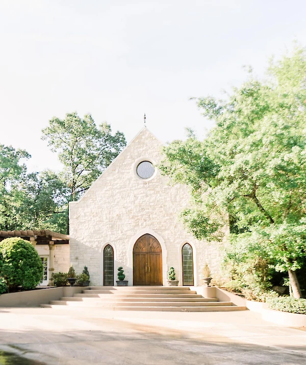 Stone chapel with arched wooden doors, narrow arched windows, and a circular window above, surrounded by mature trees and a wide staircase leading to the entrance