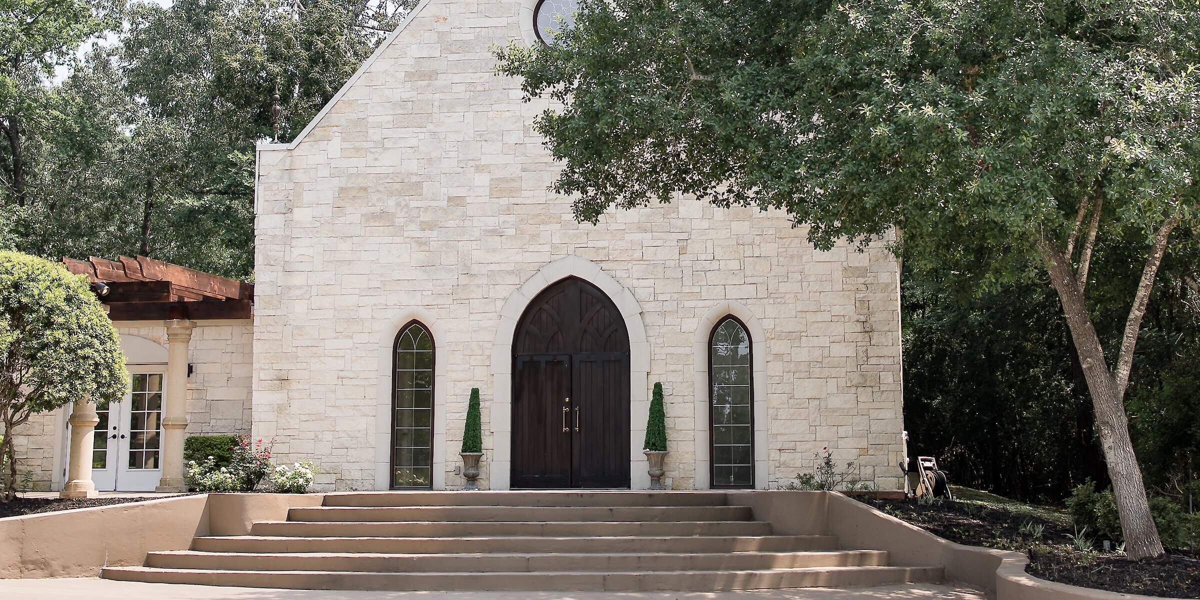Stone chapel entrance with arched wooden doors and tall narrow windows framed by light brick and greenery.