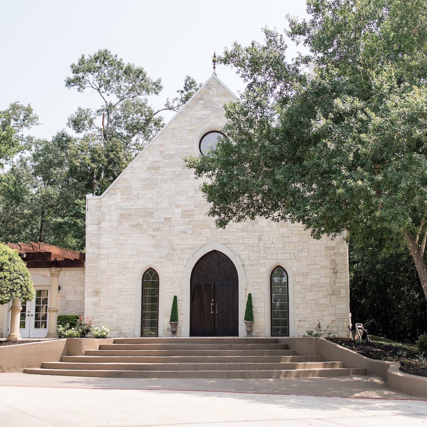 Grand stone chapel exterior with arched wooden doors, tall narrow windows, and surrounding greenery under a bright sky.