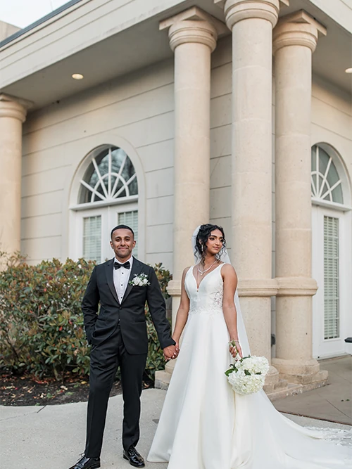 A newly married couple stands together holding a vibrant bouquet, smiling warmly at each other against a light stone wall covered in greenery.