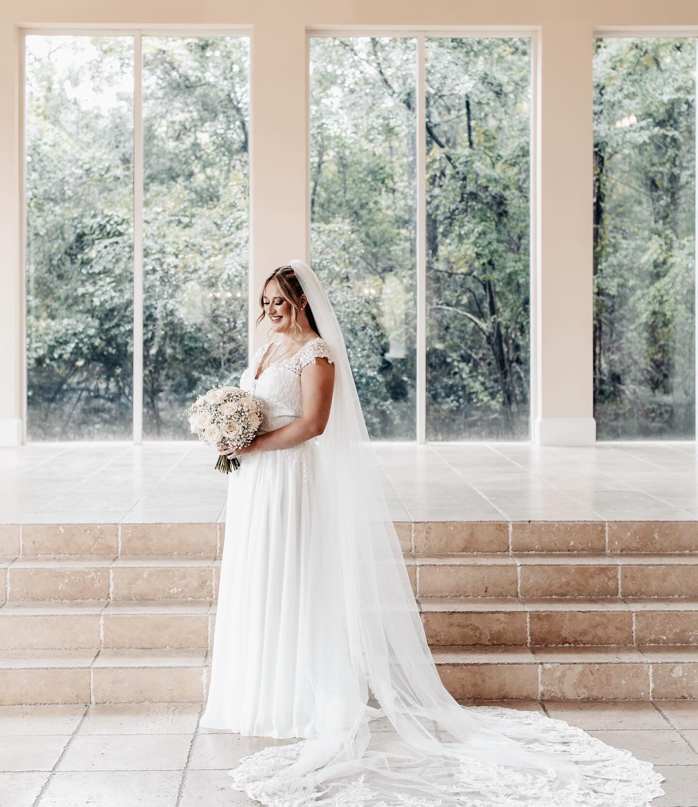 Bride standing on indoor chapel steps with a long lace train, holding a soft blush and ivory bouquet, framed by tall windows revealing a lush forest backdrop.