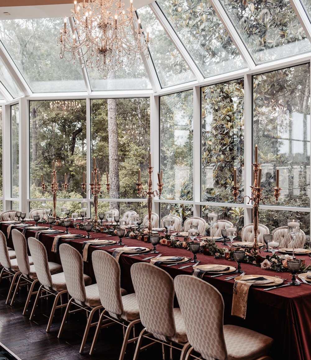 Elegant reception table setup with a long burgundy tablecloth, gold candelabras, floral garland, and place settings arranged under a crystal chandelier in a glass-walled ballroom.
