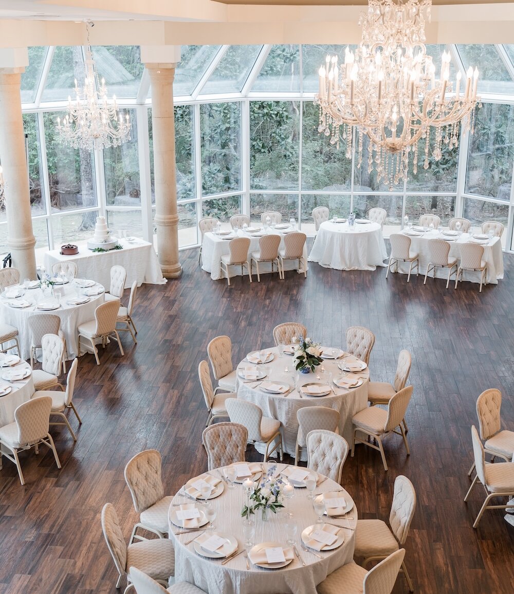 An overhead view of a wedding reception set up in a bright conservatory room with large windows, white tables, tufted chairs, and crystal chandeliers.