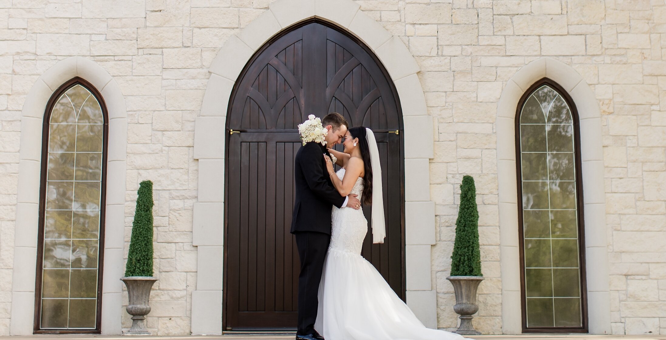 Bride and groom embrace in front of a large arched wooden door on a stone chapel, smiling with their foreheads touching.