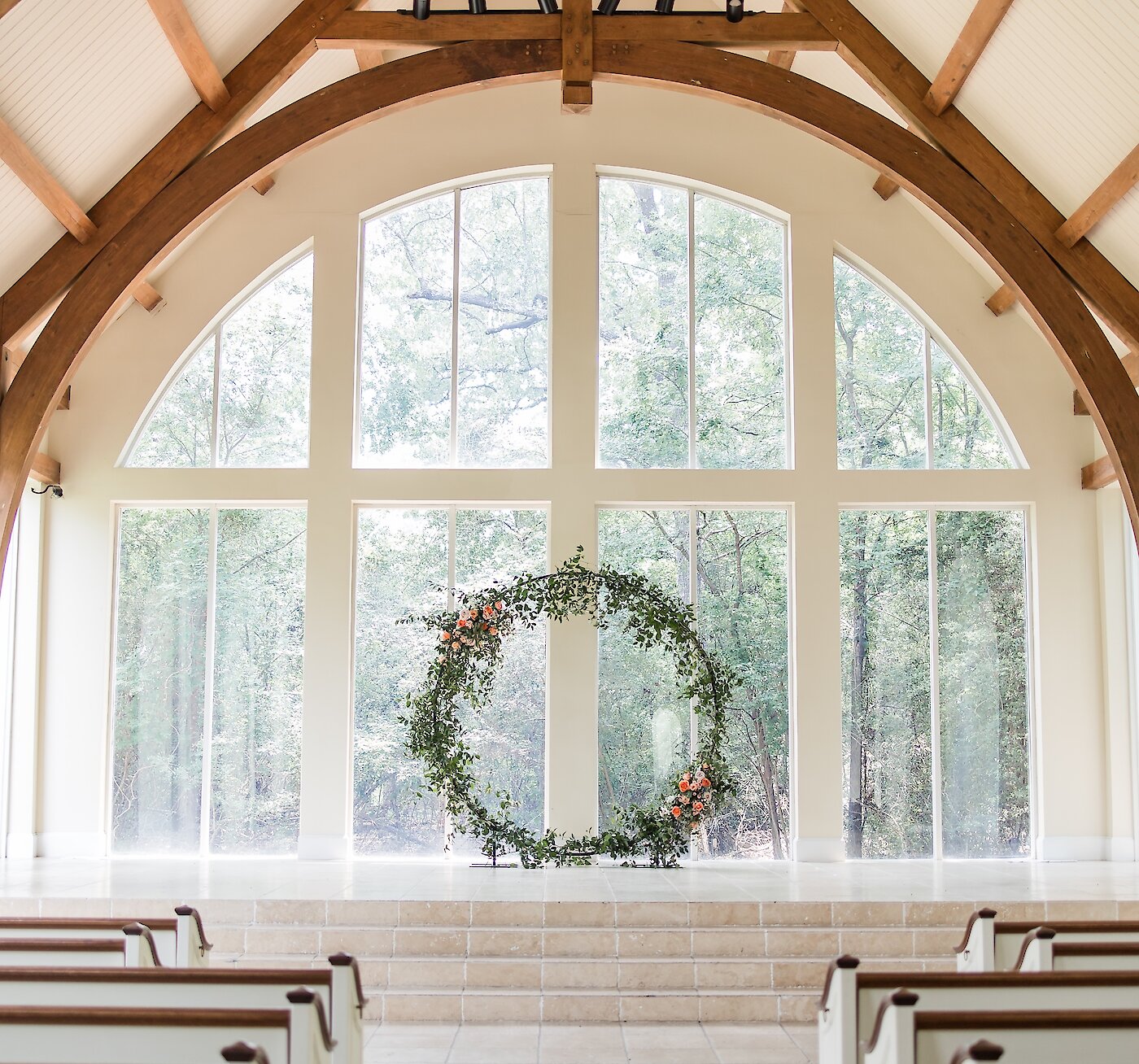 A circular greenery and floral ceremony wreath stands on the chapel stage beneath large arched windows and exposed wooden beams.
