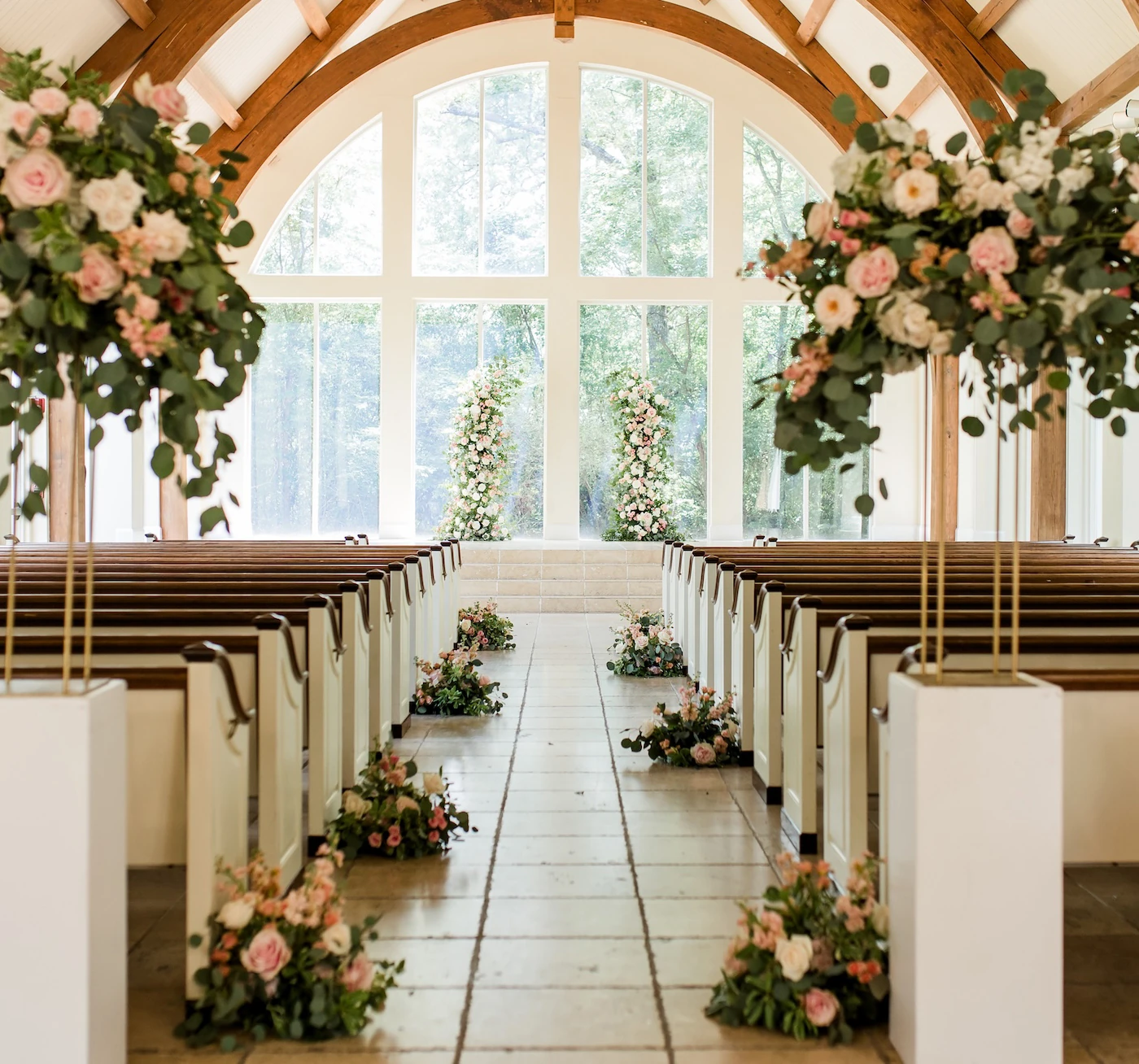 Elegant wedding chapel aisle with wooden arches, large floral arrangements of pink and white roses, and greenery leading to a bright windowed altar with matching floral pillars