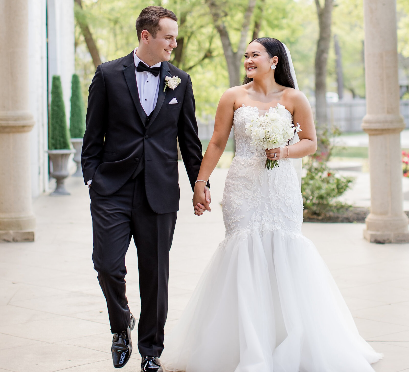 Bride and groom walking hand in hand under a wooden pergola with stone columns.