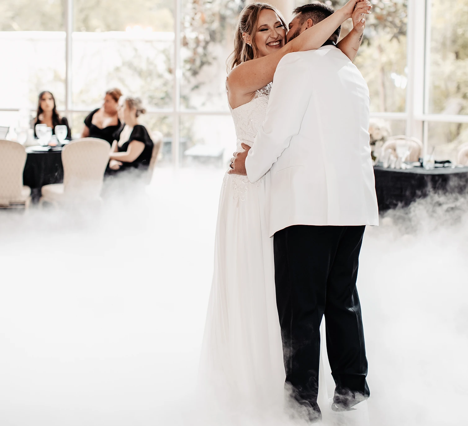 Happy married couple slow dancing surrounded by a cloud of dramatic, low-lying dry ice fog on their wedding day at an indoor venue with large windows. The bride wears a white lace dress and the groom wears a white tuxedo jacket and black pants.