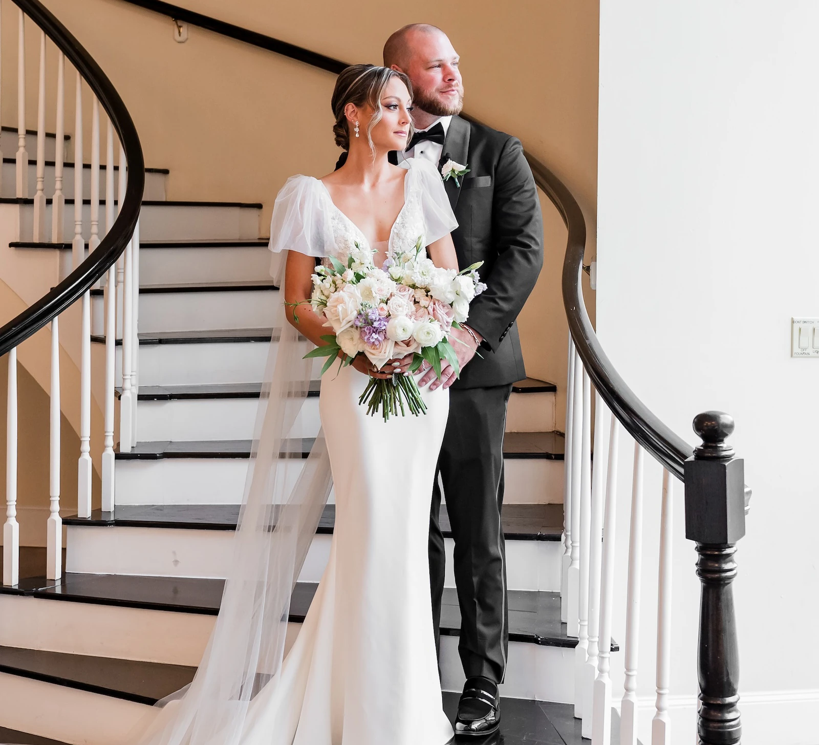 Bride and groom stand together on a grand curved staircase, the bride holding a bouquet and wearing a long veil as natural light streams in beside them.