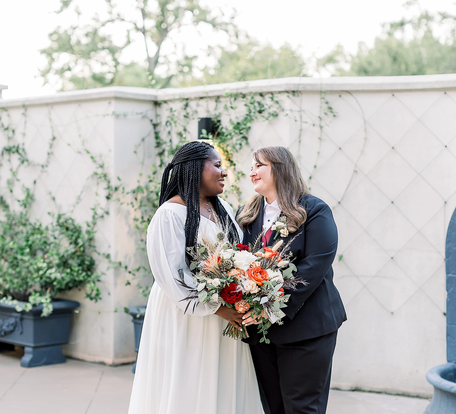 Portrait of two smiling brides posing together outdoors at a wedding venue. The bride on the left wears a white long-sleeved flowing gown and holds a large orange and white bouquet, while the bride on the right wears a black pantsuit with a black bow tie