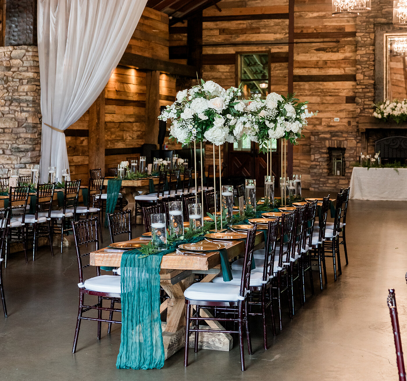 Rustic wedding reception with long wooden farm tables, emerald green runners, tall white floral centerpieces, and dark chiavari chairs inside a stone and wood lodge venue