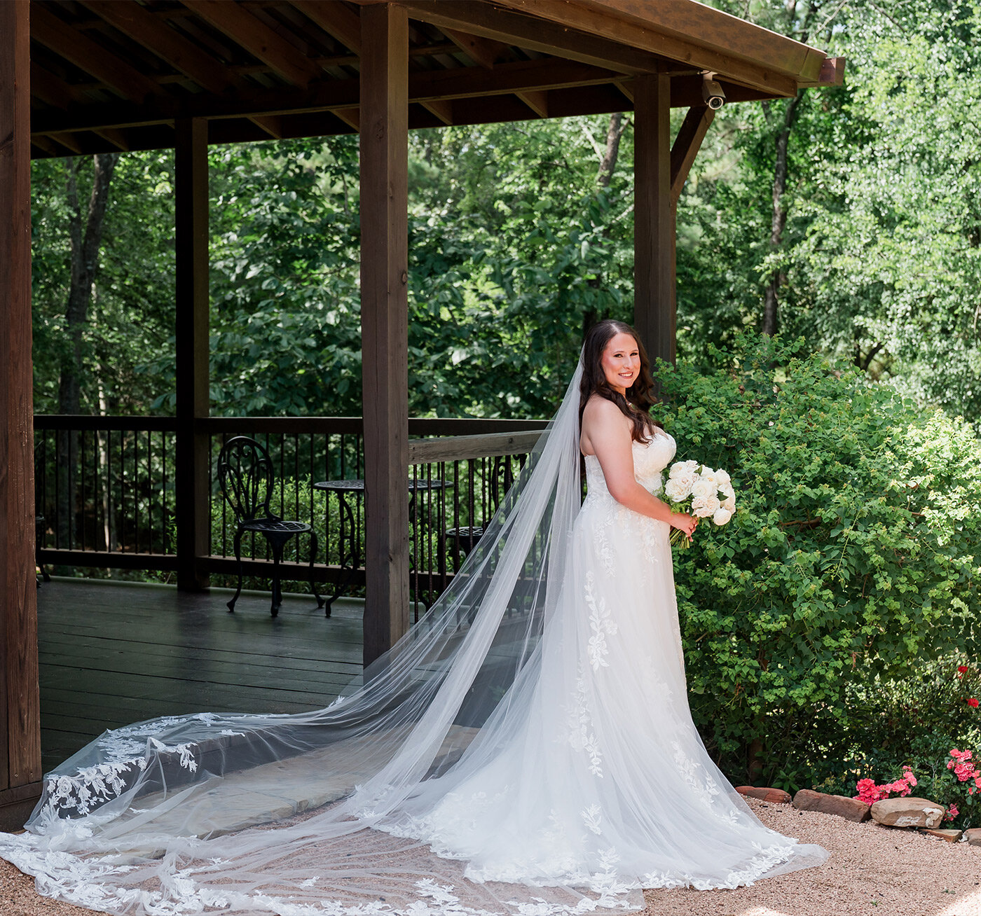 Rustic wooden wedding welcome sign with white calligraphy and floral arrangement displayed beside a large potted evergreen outside a barn-style venue
