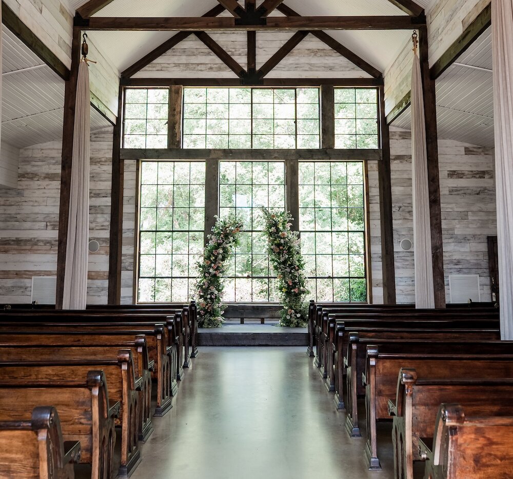 Rustic chic wedding ceremony chapel interior with a vaulted white ceiling, exposed dark wooden beams, and walls clad in whitewashed horizontal planks. The aisle is lined with traditional wooden pews leading to a large window altar decorated with colorful floral arches.