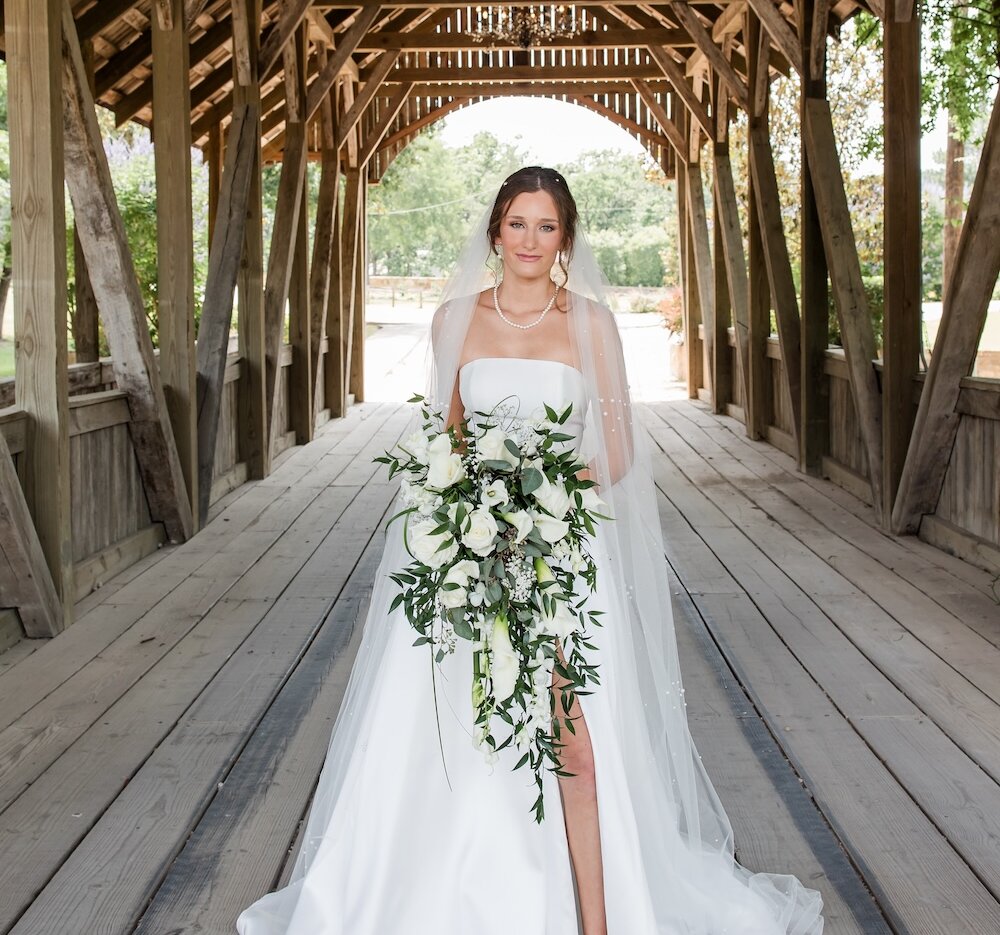 Stunning bridal portrait of the bride standing at the center of a rustic wooden covered bridge. She wears a strapless white gown with a high slit, sparkling shoes, and holds a cascading bouquet of white roses and greenery, beneath a hanging chandelier.