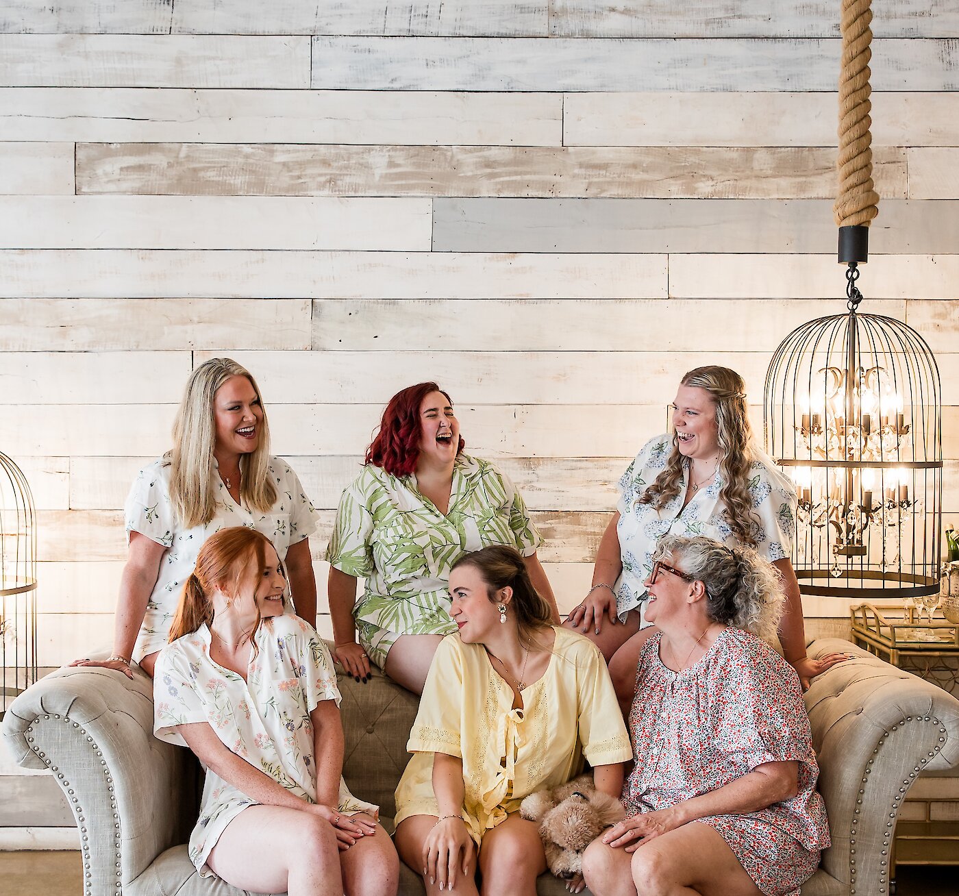 Joyful portrait of the bridal party laughing while seated on a tufted gray sofa in a rustic chic bridal suite. The women are wearing floral and patterned pajamas/robes against a whitewashed plank wall backdrop.