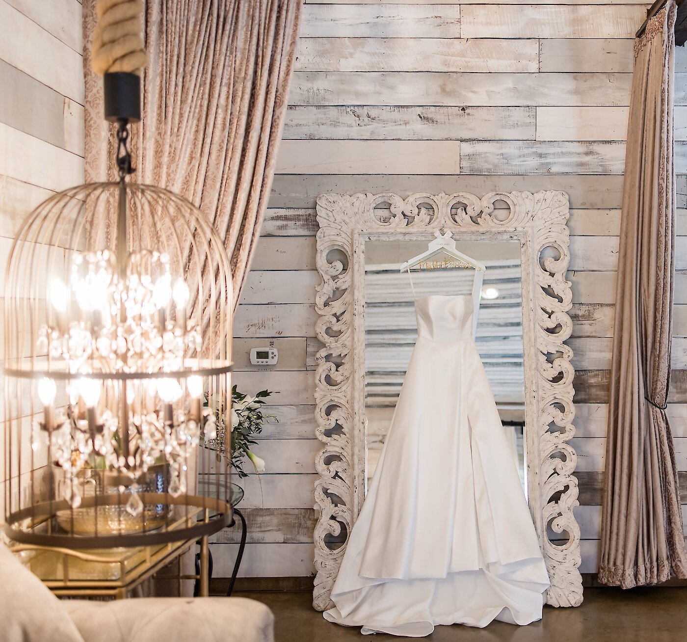 Bridal suite detail photo featuring a simple, elegant strapless white satin wedding dress hanging on a highly ornate, whitewashed wood-framed mirror. The backdrop is a whitewashed plank wall, and a rustic crystal chandelier hangs nearby.