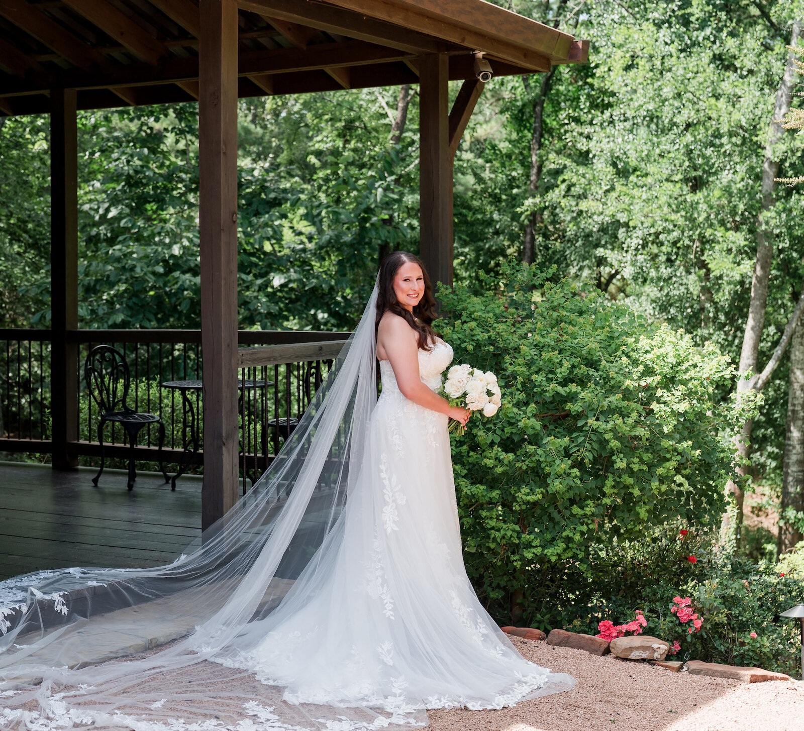 Stunning outdoor bridal portrait of the bride smiling on a rustic wooden porch, showcasing a voluminous lace gown and a dramatic cathedral-length lace-edged veil that trails onto the gravel. She holds a white rose bouquet, with lush green trees in the background.