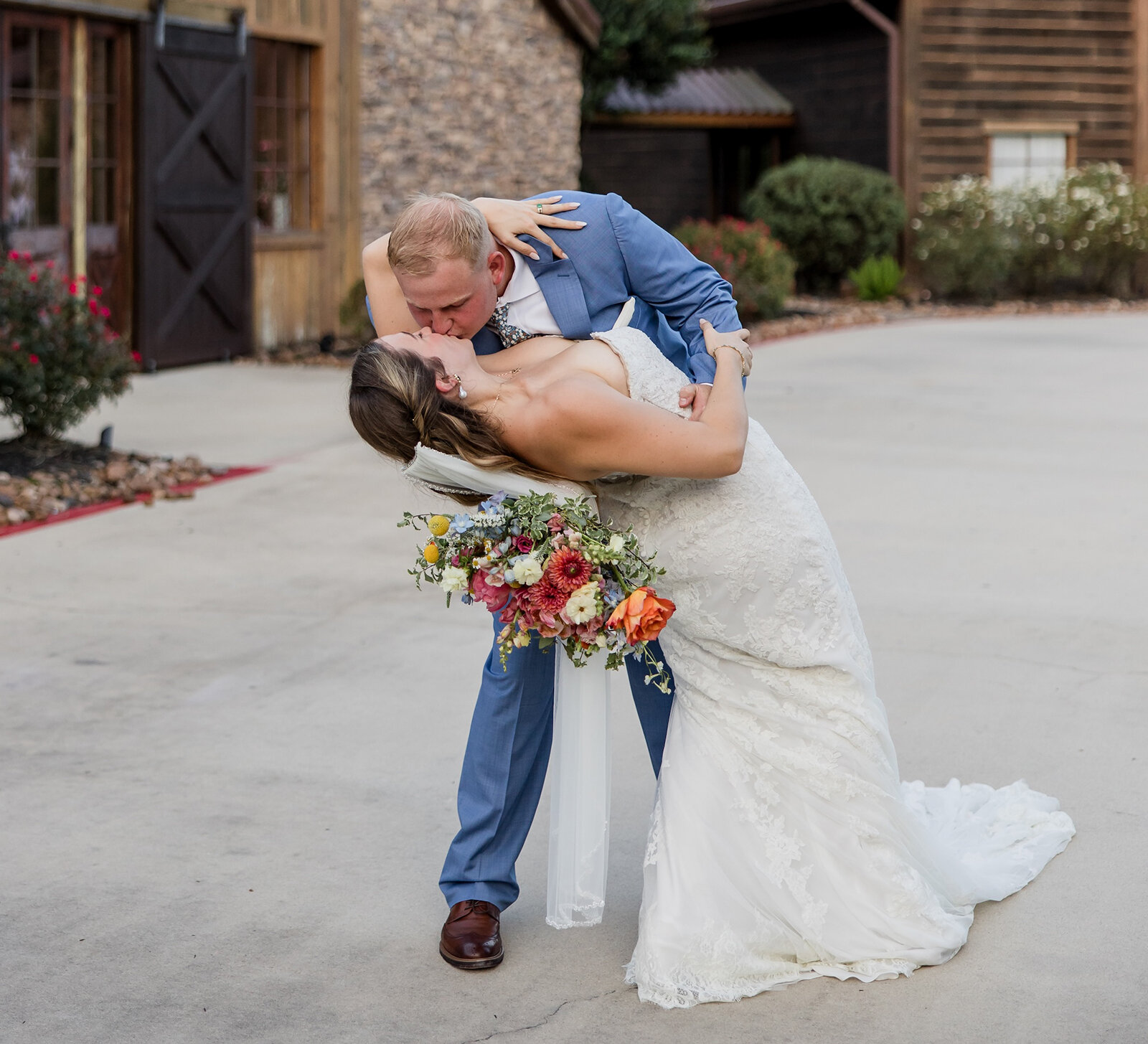Dramatic wedding kiss and dip photo outside a rustic wooden and stone barn venue. The groom wears a light blue suit, and the bride wears a strapless lace gown and holds a large, colorful bouquet.