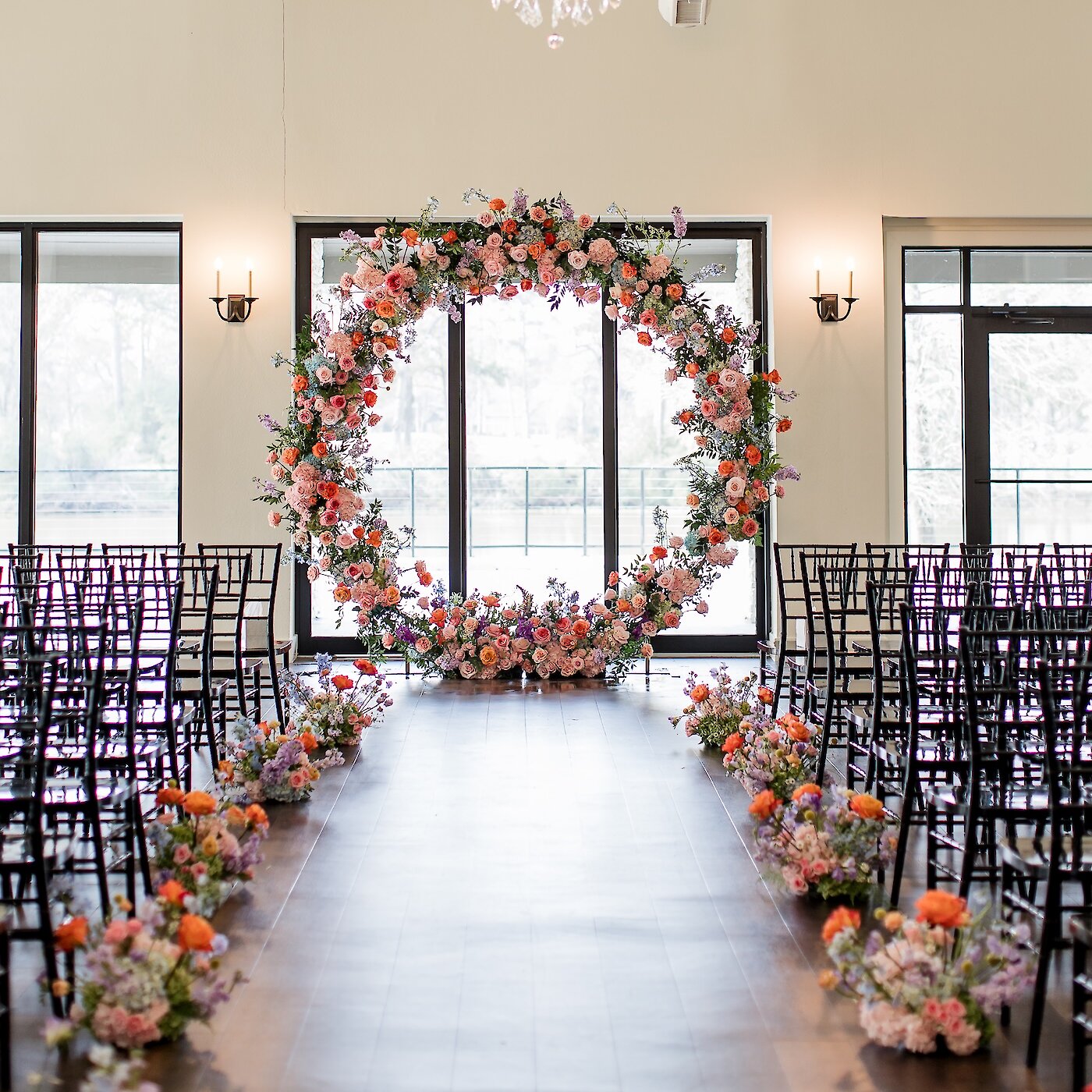Floral wedding ceremony setup with a large circular arch covered in colorful flowers and aisle arrangements beneath a sparkling crystal chandelier.