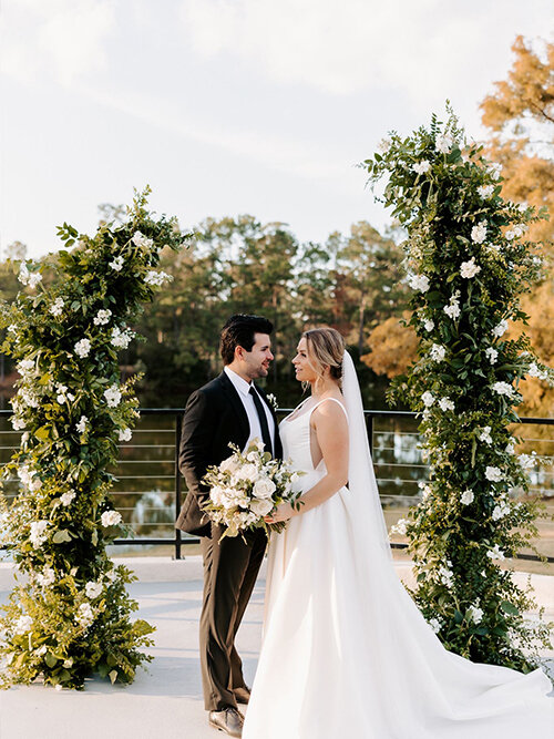 Bride and groom pose indoors, with the bride holding a white floral bouquet and her long veil flowing across the floor as the groom stands beside her in a navy suit.