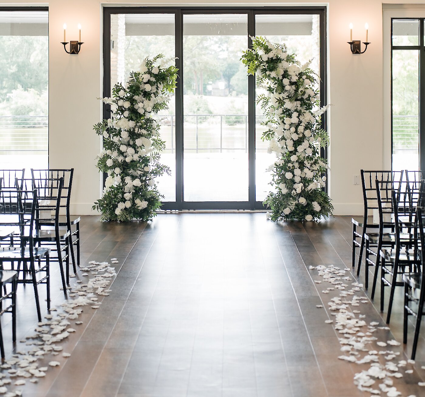 An elegant indoor wedding ceremony aisle with black chairs, white petals, a crystal chandelier, and a curved white and green floral arch framing the doors to a lake.