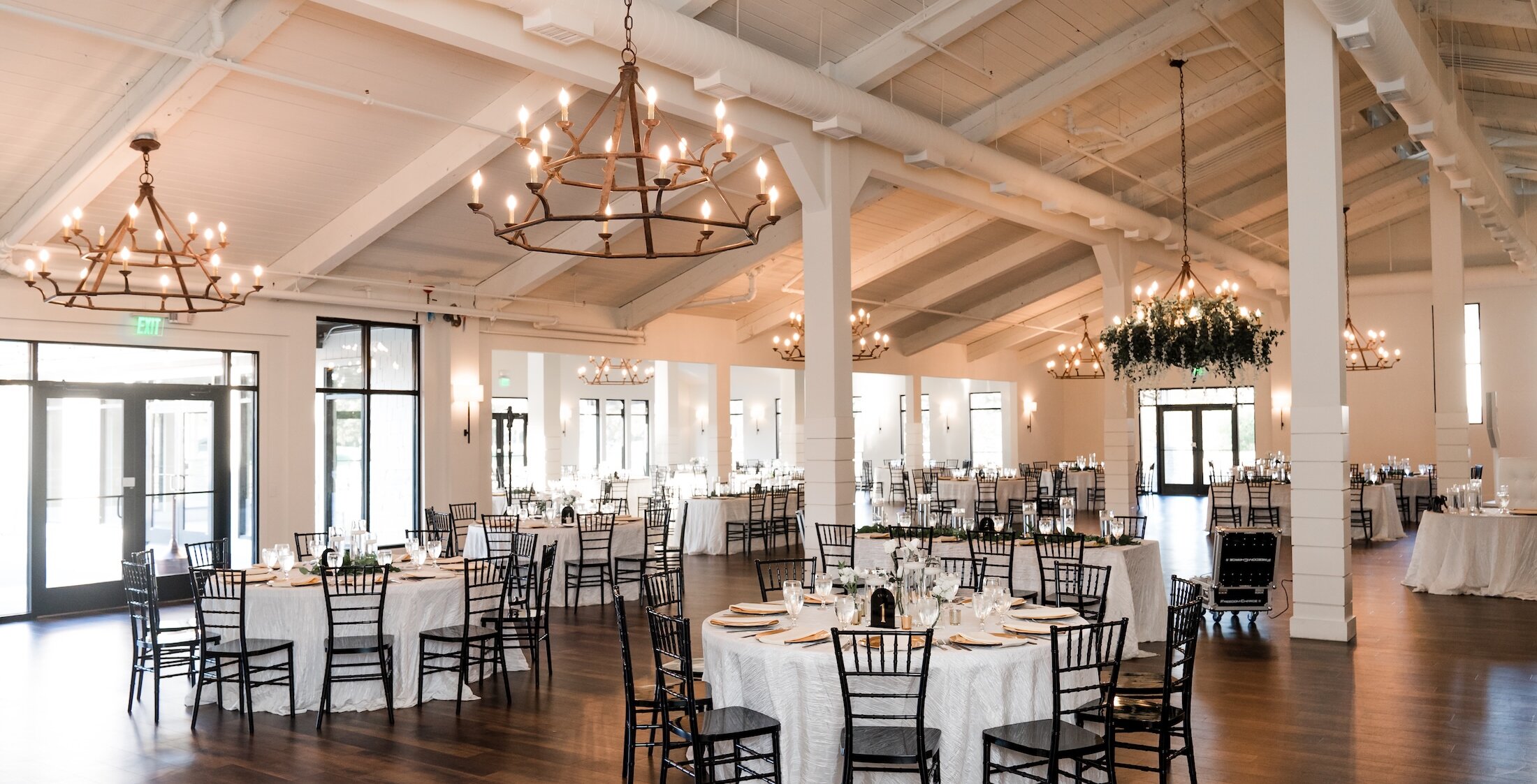 Spacious wedding reception hall with round tables set with white linens, black chiavari chairs, elegant chandeliers, and greenery accents throughout the bright, open space.