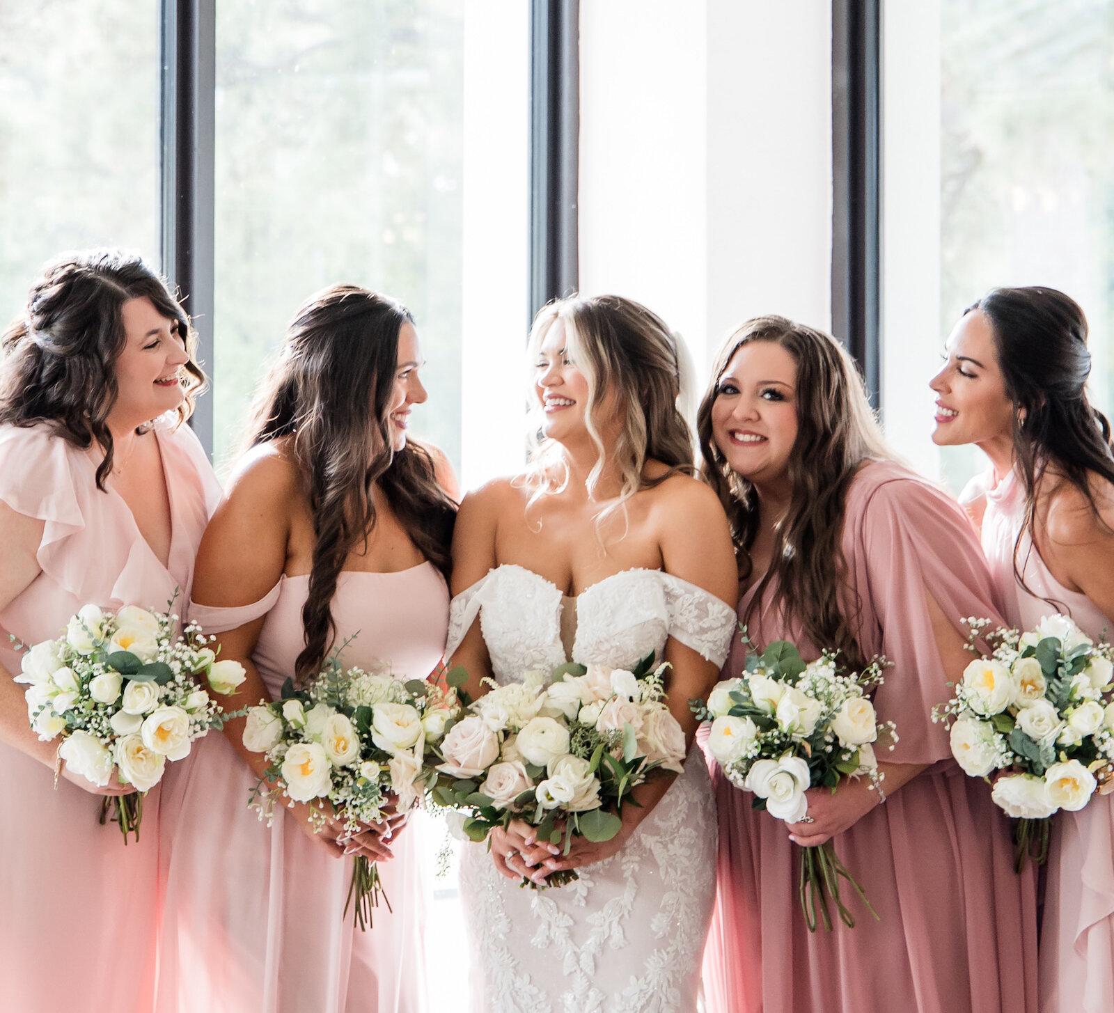 Bride standing with her bridesmaids in blush pink dresses, all holding white and pastel bouquets, smiling together in front of tall bright windows.