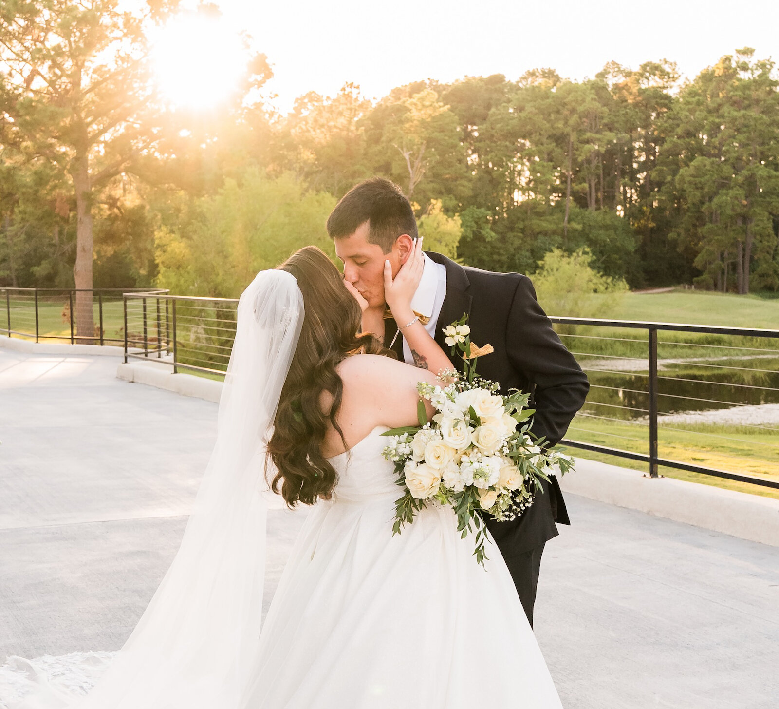 Bride and groom sharing a kiss at sunset on an outdoor terrace, with the bride holding a white floral bouquet and her long veil flowing behind her.