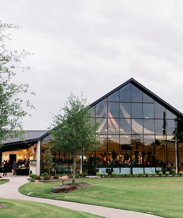 Exterior of a modern white chapel wedding venue with vertical siding, a central arched black-framed door, and a small bell tower. The complex is set on a manicured green lawn under a blue sky, next to a contemporary glass building structure.