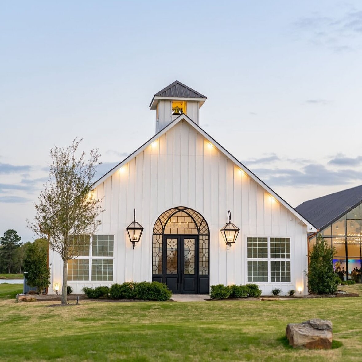 Exterior of a modern white chapel wedding venue with vertical board siding, a small bell tower, and a dramatic arched black iron door. The building is illuminated by exterior lights at dusk, set on a green lawn next to a contemporary glass-front structure.