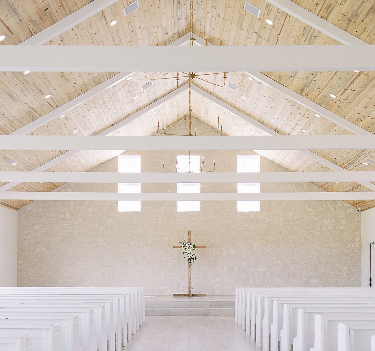 The bright interior of a farmhouse-style wedding chapel with a vaulted wood ceiling, white stone walls, white pews, and a floral-decorated wooden cross.