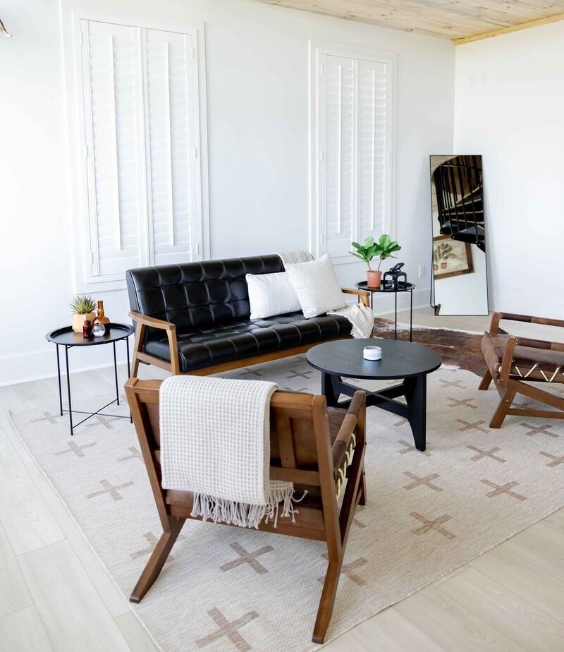 Modern rustic interior lounge featuring a black leather sofa, mid-century modern wooden armchairs, and a beige patterned rug. The room has white walls, plantation shutters, and a slatted wood plank ceiling with a black chandelier.