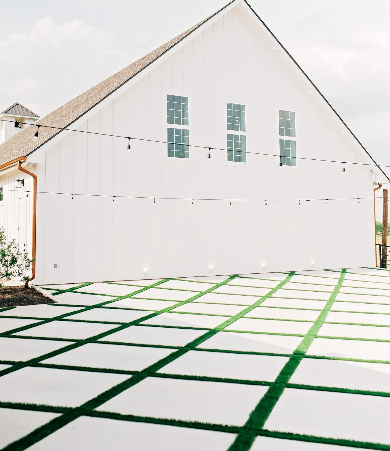 The exterior of a white modern farmhouse venue with three upstairs windows and a patio featuring a checkerboard pattern of white paving and green turf.