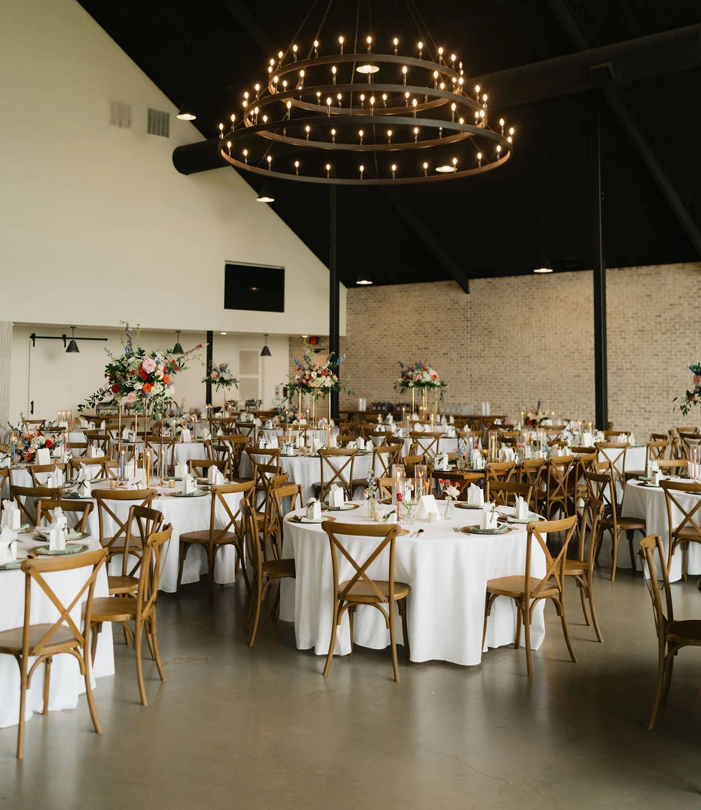 Rustic modern wedding reception hall featuring a high vaulted black ceiling with a large circular wagon wheel chandelier. Round tables are set with white linens, light wood cross-back chairs, and vibrant floral centerpieces with red and blue accents, set against a light brick wall.