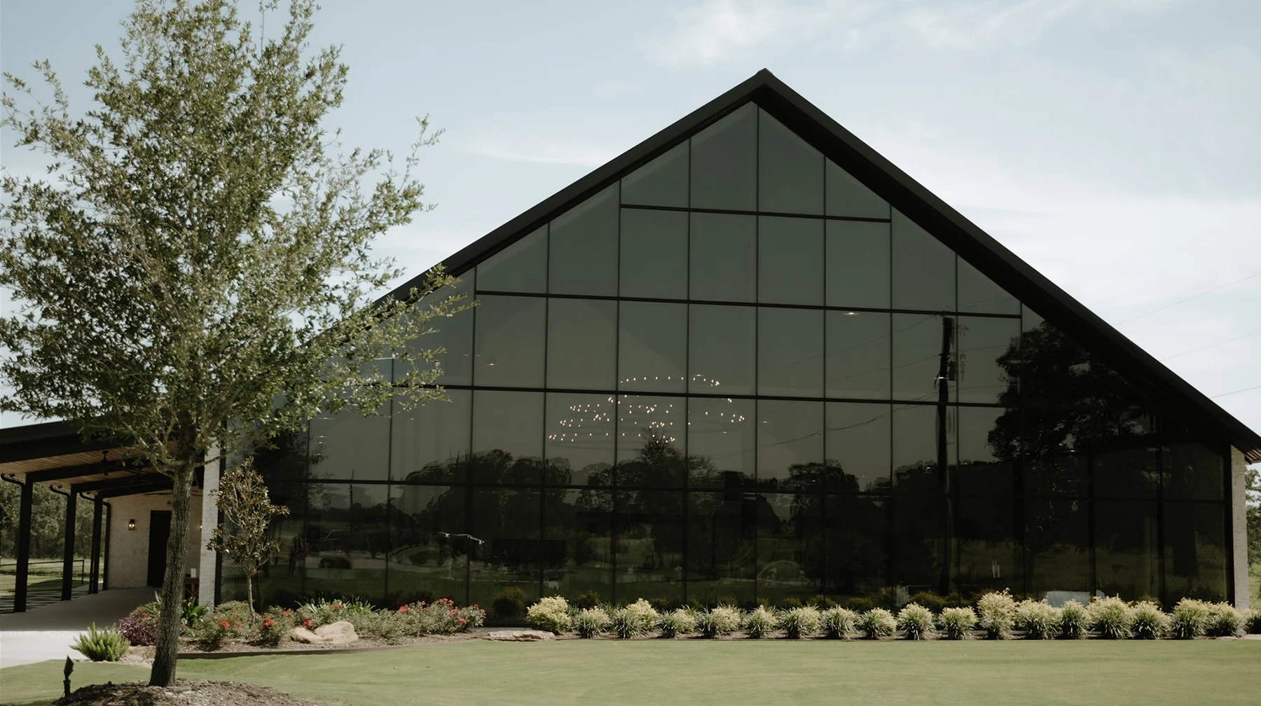 Exterior of a modern glass-front wedding venue with a black A-frame facade visible through the windows at dusk. The interior is brightly lit for a reception, and a curved walkway leads across the manicured lawn toward the entrance.