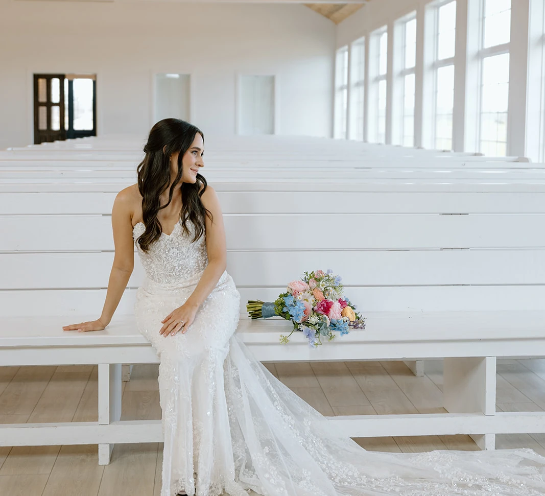 Bride in a lace wedding gown sitting on a white wooden pew with a pastel floral bouquet in a bright chapel with a wood-plank ceiling