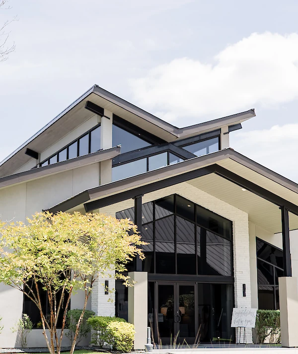Modern wedding venue exterior with sharp angular rooflines, large black-framed windows, and landscaped greenery on a sunny day.