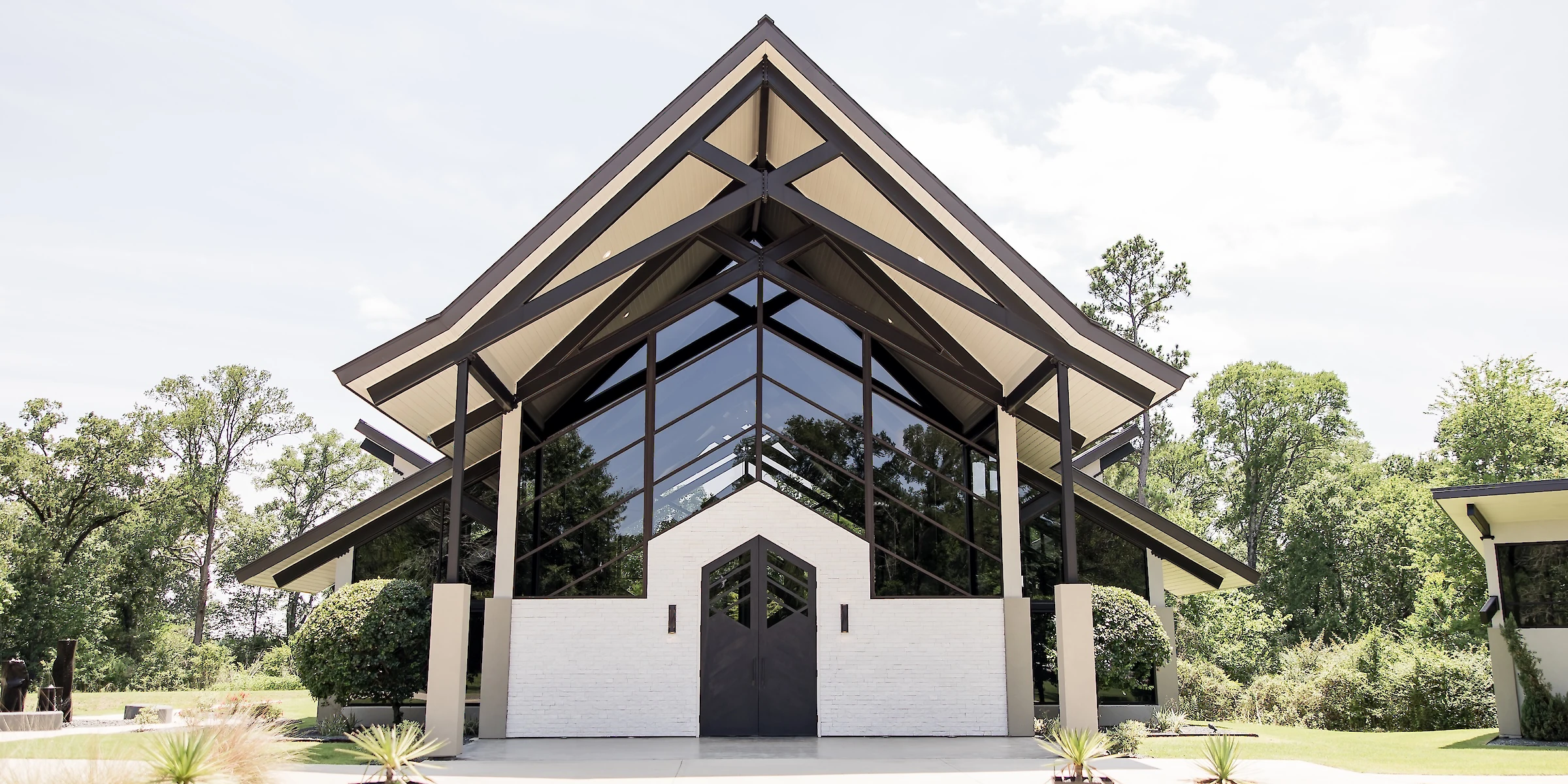 Modern glass-front wedding chapel with striking angular rooflines, surrounded by lush green trees under a bright, clear sky.