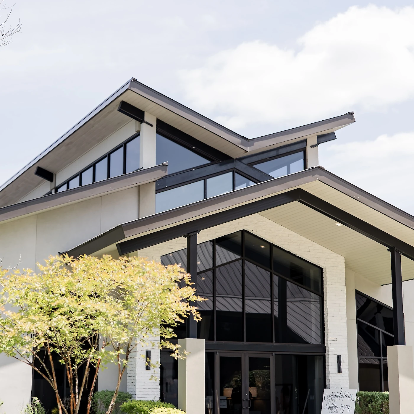 Modern wedding venue exterior featuring sharp architectural lines, expansive black-framed windows, and landscaped greenery on a sunny day.