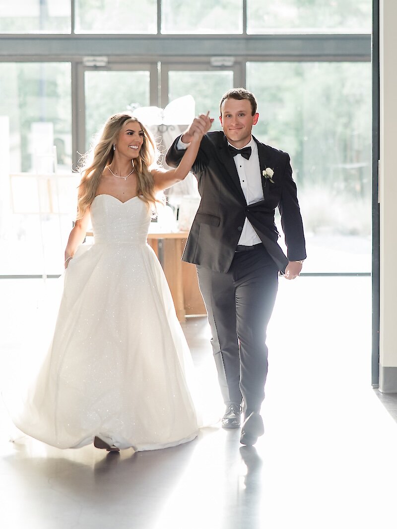 Bride and groom make a joyful grand entrance into their wedding reception, holding hands and smiling as sunlight pours in from behind them.
